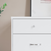 A close-up of the Child Craft Soho 3-Drawer Dresser in White/Natural shows its distinct knobs and handles, placed beside a green plant with a framed picture partially visible on the wall above.