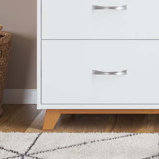 A close-up of the Child Craft Soho 3-Drawer Dresser in White/Natural, featuring silver handles and wooden legs, stands next to a woven basket on a wooden floor with a patterned rug.
