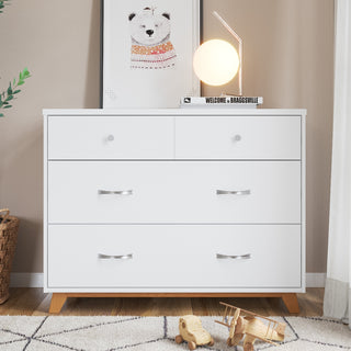 The Child Craft Soho 3-Drawer Dresser in White/Natural stands on a rug with a framed bear print, round lamp, and book on top. Toy airplanes and a car are nearby, with a basket and curtain in the background.