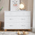 The Child Craft Soho 3-Drawer Dresser in White/Natural stands on a rug with a framed bear print, round lamp, and book on top. Toy airplanes and a car are nearby, with a basket and curtain in the background.