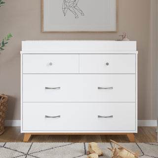 The Child Craft Soho 3-Drawer Dresser in White/Natural features sleek light wood legs and silver handles, shown on a nursery rug beneath a framed geometric animal print, with a basket and toys placed nearby.