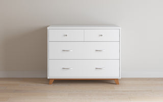 The Child Craft Soho 3-Drawer Dresser in White/Natural, featuring minimalist handles and short wooden legs, stands against a light beige wall on a wooden floor.