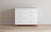 The Child Craft Soho 3-Drawer Dresser in White/Natural, featuring minimalist handles and short wooden legs, stands against a light beige wall on a wooden floor.