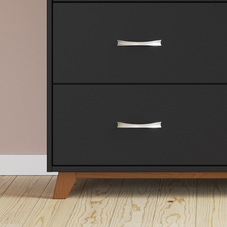 The lower half of the Child Craft Soho 3-Drawer Dresser in Ebony, featuring silver handles and wooden legs, is shown standing on a light wood floor against a beige wall.