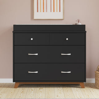 The Child Craft Soho 3-Drawer Dresser in Ebony features silver handles and wooden legs, set against a pale pink wall with a changing pad and soap dispenser on top. Above hangs abstract art, while a woven basket rests on the floor to the right.