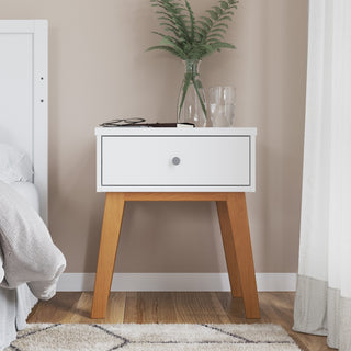 The Child Craft Soho Nightstand in White/Natural stands beside a bed, topped with a potted fern, glass of water, eyeglasses, and a book. A nearby rug and curtain are partially visible.