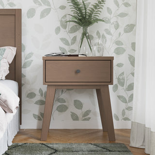 The Child Craft Soho Nightstand in Dusty Heather stands beside a bed, topped with a glass of water, a book, eyeglasses, and a vase of ferns, against leafy green-patterned wallpaper.