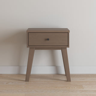 The Child Craft Soho Nightstand in Dusty Heather features a single drawer with a round black knob and four angled legs, making it perfect as nursery furniture or a kid’s nightstand against a light wall on hardwood floors.