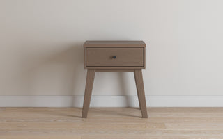 The Child Craft Soho Nightstand in Dusty Heather features one drawer and four angled legs, styled in mid-century modern design, and sits against a white wall on a light wood floor.