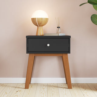 The Soho Nightstand by Child Craft in ebony, with wooden legs and a black drawer, stands against a light pink wall. On top are a round wooden lamp, small book, glass reed diffuser, and a green plant partially visible on the right.