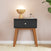 The Soho Nightstand by Child Craft in ebony, with wooden legs and a black drawer, stands against a light pink wall. On top are a round wooden lamp, small book, glass reed diffuser, and a green plant partially visible on the right.