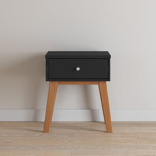 The Child Craft Soho Nightstand in Ebony features a black drawer with a round silver knob and stands on four slanted wooden legs, set against a plain white wall and light wood flooring.