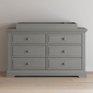 A Child Craft dresser featuring the F01792 Universal Changing Table Topper in Lunar Gray, six drawers with round knobs, rests against a plain white wall on a light wood floor.