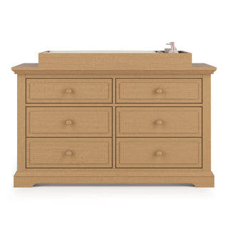 The Child Craft Legacy Changing Table Topper in Biscotti is placed on a wooden six-drawer dresser with round knobs, featuring a mat and a bottle of lotion or soap on the right side.