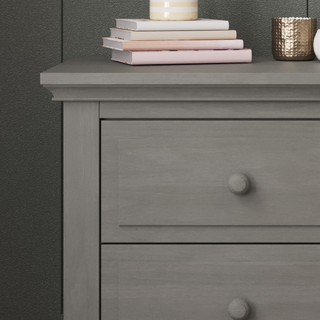 A Child Craft Jordyn Nightstand in lunar gray with crown molding and baby-safe finishes sits against a dark wall, topped with pastel books, a white and gold striped candle, and a textured metallic gold cup.