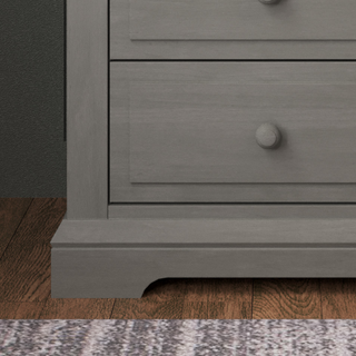 A close-up of the Child Craft Jordyn Nightstand in Lunar Gray shows its baby-safe finish, two drawers with round knobs, and carved base, standing on a wooden floor beside a patterned rug.