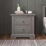 A Child Craft Jordyn Nightstand, Ready-to-Assemble in Lunar Gray, with two drawers stands on a wooden floor. It features elegant crown molding and baby-safe finishes for style and peace of mind. On top are books, a pencil jar, cup, and décor.