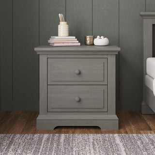A Child Craft Jordyn Nightstand, Ready-to-Assemble in Lunar Gray, with two drawers stands on a wooden floor. It features elegant crown molding and baby-safe finishes for style and peace of mind. On top are books, a pencil jar, cup, and décor.