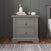 A Child Craft Jordyn Nightstand, Ready-to-Assemble in Lunar Gray, with two drawers stands on a wooden floor. It features elegant crown molding and baby-safe finishes for style and peace of mind. On top are books, a pencil jar, cup, and décor.