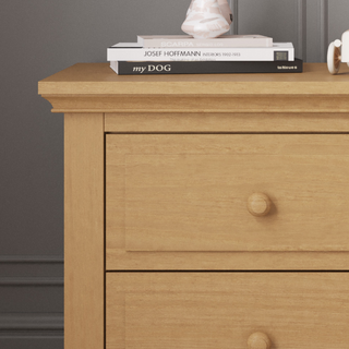 A Child Craft Jordyn Nightstand, Ready-to-Assemble in Biscotti, features three drawers. On top are two stacked books (one titled "my DOG"), a white mug, and a decorative object against a dark gray wall with crown molding.