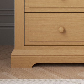 A close-up of the Child Craft Jordyn Nightstand, Ready-to-Assemble in Biscotti, displays two drawers with round knobs and crown molding. The nightstand is on a wooden floor, partly beside a dark gray wall.