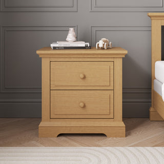 A Child Craft Jordyn Nightstand, Ready-to-Assemble in Biscotti, with two drawers is set against a gray paneled wall with crown molding. On top are stacked books, a small vase, and a wooden toy; a matching bed frame appears on the right.