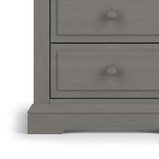 Close-up of the bottom left corner of the Child Craft Jordyn Nightstand in Lunar Gray, showing two drawers with round knobs. The nightstand features a simple, classic design, baby-safe finishes, and a solid base. Ready-to-assemble.