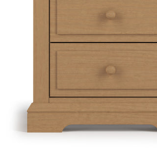 A close-up of the lower left corner of the Child Craft Jordyn Nightstand, Ready-to-Assemble in Biscotti, featuring two drawers with round knobs, a curved base, and crown molding—ideal as a nursery nightstand.