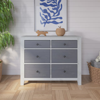 The Child Craft Ocean Grove Double Dresser in White/Gray features six dark gray drawers and stands on a wood floor, making it ideal nursery furniture. Accessories like a plant and art add nautical farmhouse charm.