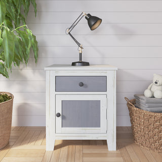 The Child Craft Ocean Grove Nightstand in white/gray stands on a wood floor with a black desk lamp on top, a green plant in a basket to the left, and a basket holding towels and a teddy bear to the right.