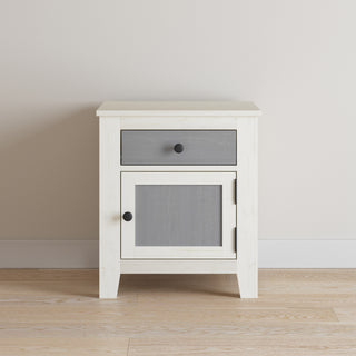 The Child Craft Ocean Grove Nightstand in White/Gray features a single gray drawer and cabinet door with black knobs, set on a light wood floor against a pale wall.