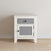 The Child Craft Ocean Grove Nightstand in White/Gray features a single gray drawer and cabinet door with black knobs, set on a light wood floor against a pale wall.