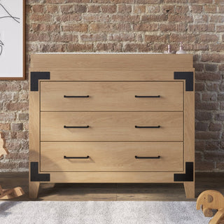 A modern wooden dresser with black metal accents stands before a brick wall, topped with the Child Craft F01792 Universal Changing Table Topper in Nutmeg. Part of framed artwork and wooden toys are visible nearby.