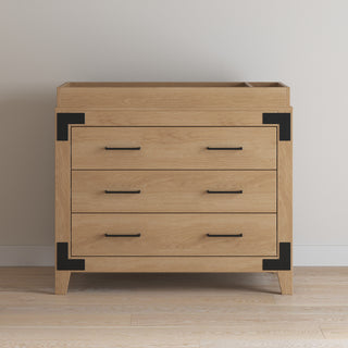 The Child Craft Lucas Dresser in Nutmeg, featuring three drawers with black metal handles and corner accents, stands on a wooden floor against a plain white wall.