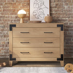 The Child Craft Lucas Dresser in Nutmeg, featuring three drawers with black handles, is set against an exposed brick wall. On top sit a round lamp, decorative bowl, small vase, and abstract artwork. A portion of a rug appears below.