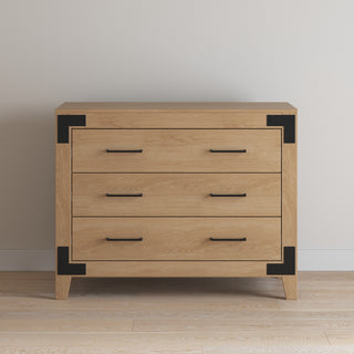 The Lucas Dresser by Child Craft in Nutmeg features three drawers with black metal handles and corner brackets. It stands on four short legs and is shown against a white wall on a light wood floor.