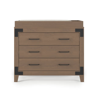 The Child Craft Lucas Dresser in Dusty Heather features three drawers with double black handles, black metal corner accents, short angled legs, and a top surface that's slightly raised at the edges.