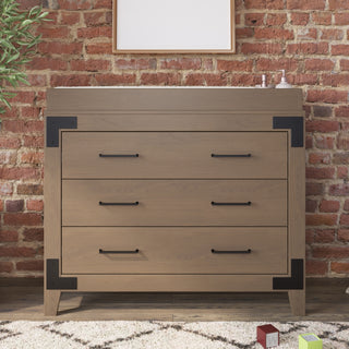The Child Craft Lucas Dresser in Dusty Heather, featuring three drawers and black metal corner accents, stands against a red brick wall with decor items on top, a leafy plant to the left, and blocks with a rug in front.