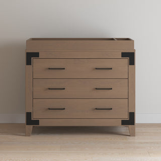 The Lucas Dresser in Dusty Heather by Child Craft, featuring three drawers with black metal handles and black corner brackets, stands against a light wall on a wooden floor.