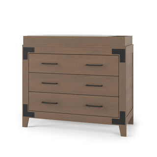 The Child Craft Lucas Dresser in Dusty Heather features three drawers with black metal handles and corner brackets, a rustic design, and angled legs, set against a white background.