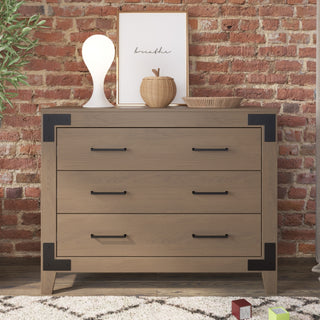The Child Craft Lucas Dresser in Dusty Heather, with three drawers and black metal accents, stands against a brick wall. On top sit a white lamp, wicker basket, framed "breathe" print, and tray; toys are scattered nearby.