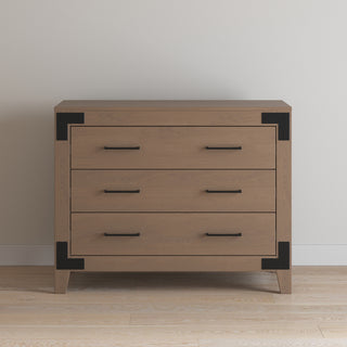 The Child Craft Lucas Dresser in Dusty Heather features three drawers, black metal corner accents, and matching black handles. It stands on a light wood floor against a plain off-white wall.