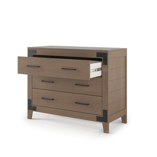 The Child Craft Lucas Dresser in Dusty Heather features three drawers with black metal handles and metal corner accents. The top drawer is open, showing a neatly stacked item inside. The dresser stands on four short legs.