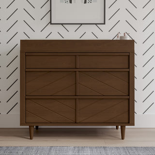 The Child Craft Surrey Hill 3 Drawer Dresser in Toasted Chestnut, featuring angled legs and geometric patterns, stands against a white wall with black diagonal lines. Framed art and decor items are arranged on top.
