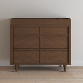 The Child Craft F01792 Universal Dressing Kit in Toasted Chestnut, featuring three drawers with geometric fronts and tapered legs, stands against a light wall on a wood floor and includes a convenient changing table topper for added function.
