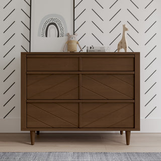 The Child Craft Surrey Hill 3 Drawer Dresser in Toasted Chestnut stands before a white wall with black lines, topped with a framed rainbow print, plush toy, wooden giraffe, camera, and a decorative box.