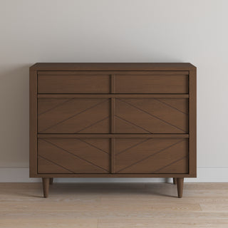 The Child Craft Surrey Hill 3 Drawer Dresser in Toasted Chestnut features a geometric patterned front and angled legs, and stands against a light-colored wall on a wooden floor.