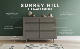 The Child Craft Surrey Hill 3 Drawer Dresser in Lunar Gray stands against a green wall, showcasing diamond-patterned fronts, built-in handles, angled legs, and top decor that accentuates its spacious drawers and elegant finish.