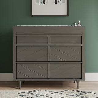 A Child Craft Surrey Hill 3 Drawer Dresser in Lunar Gray stands against a green wall, topped with soap dispensers and framed abstract artwork above. The patterned rug below completes this stylish nursery furniture setup.