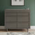 A Child Craft Surrey Hill 3 Drawer Dresser in Lunar Gray stands against a green wall, topped with soap dispensers and framed abstract artwork above. The patterned rug below completes this stylish nursery furniture setup.